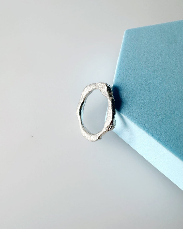 Handmade recycled sterling silver ring band with an organic, molten texture and irregular shape, photographed from above leaning against a blue prop.