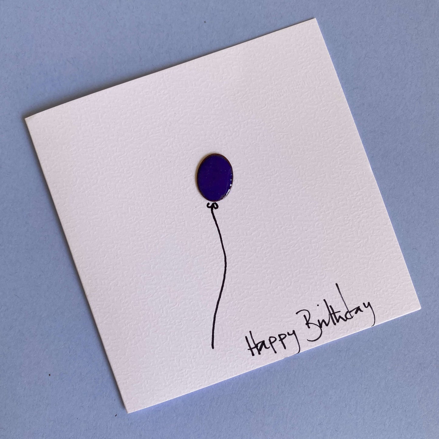 blue enamel balloon on the front of white greeting card with Happy Birthday handwritten text
